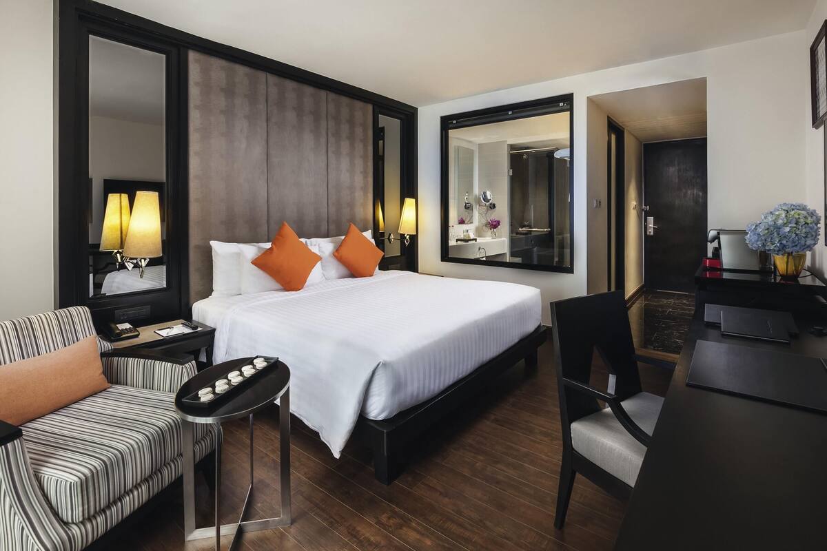 A hotel room with a bed, a desk, a chair, a sofa, a mirror, and a bathroom visible through the door.