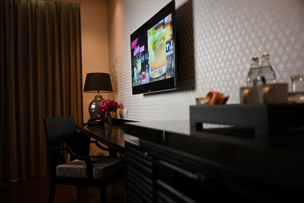 Habitación de hotel con televisión montada en la pared, una mesa con lámpara, una silla y un jarrón decorativo con flores.