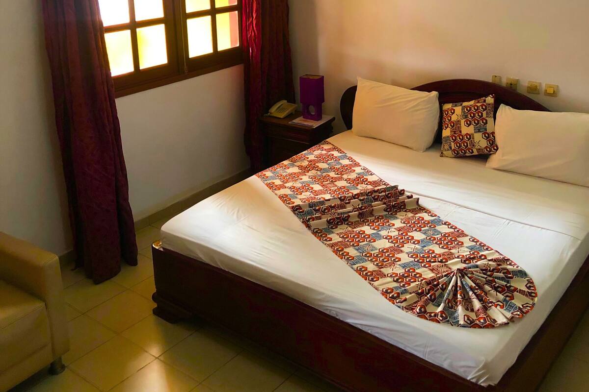 A hotel room with a large bed, a bedside table, a lamp, a window with curtains, and a wall-mounted light.