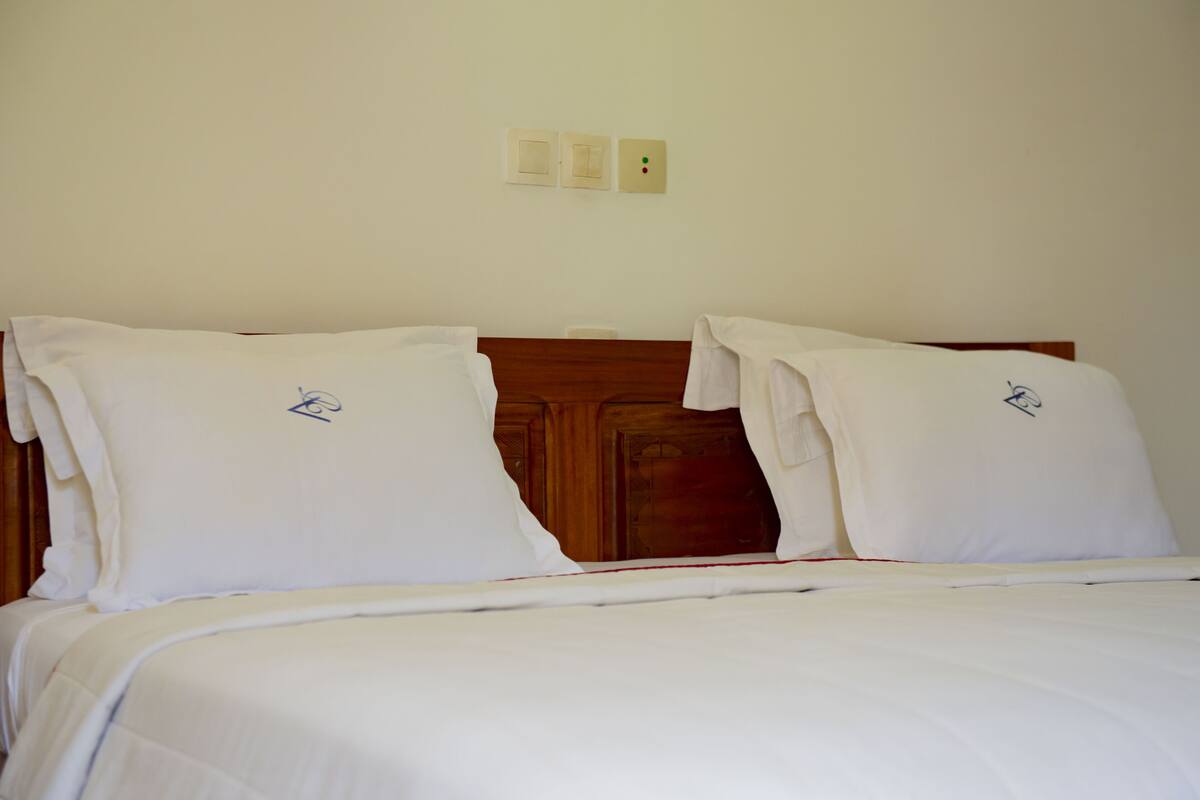 A neatly made bed with white linens and pillows, a wooden headboard, and a plain wall with two light switches.