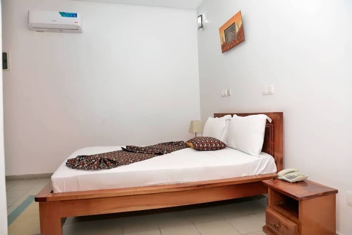 A single bed with a wooden frame, white bedding, and a patterned blanket. There is a bedside table with a lamp and a telephone. An air conditioning unit is mounted on the wall above the bed.