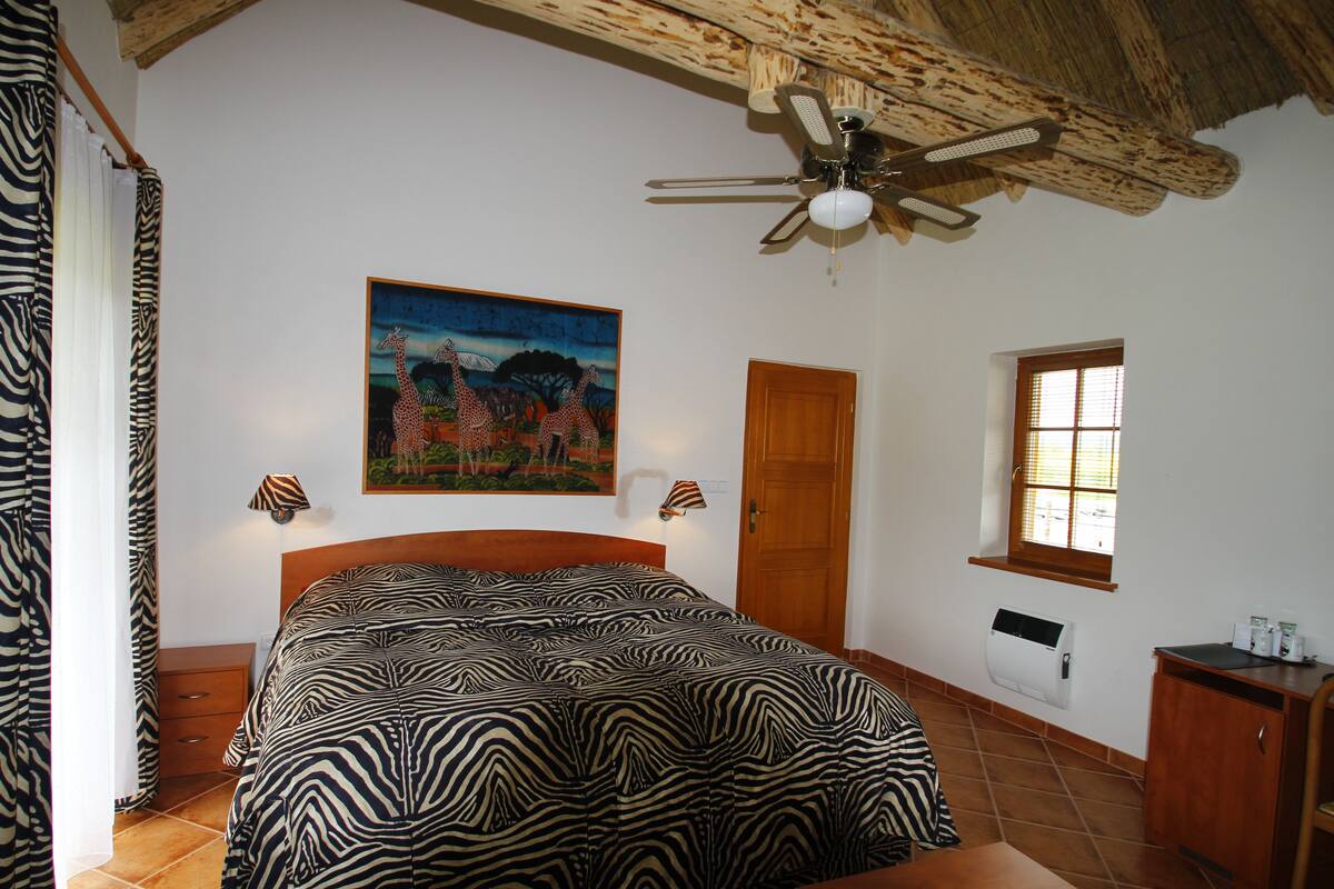 A bedroom with a zebra-patterned bedspread, a ceiling fan, a painting of giraffes, a wooden door, a window, and a small cabinet.