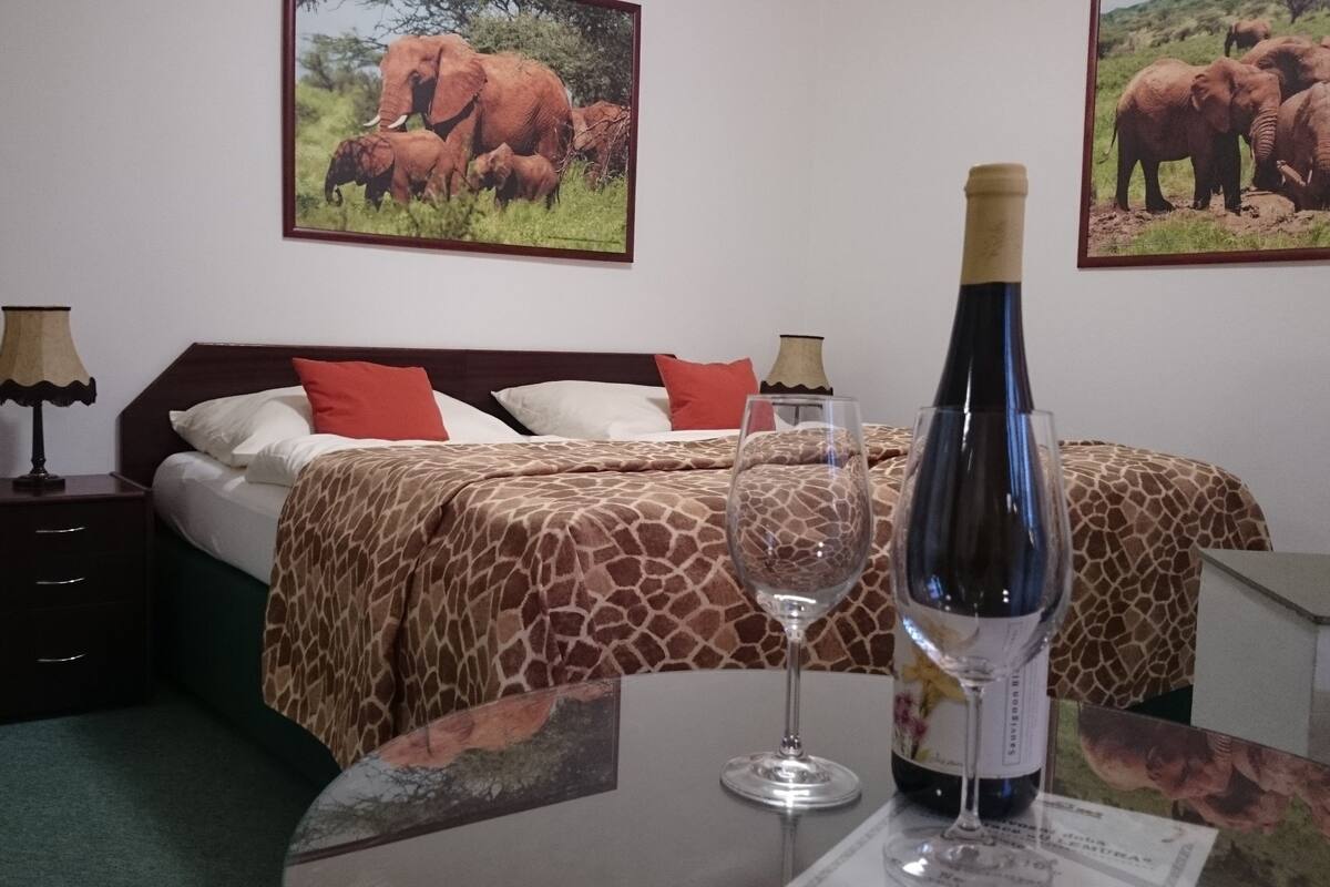 A hotel room with a bed, a nightstand, a lamp, a bottle of wine, and two framed pictures of elephants on the wall.