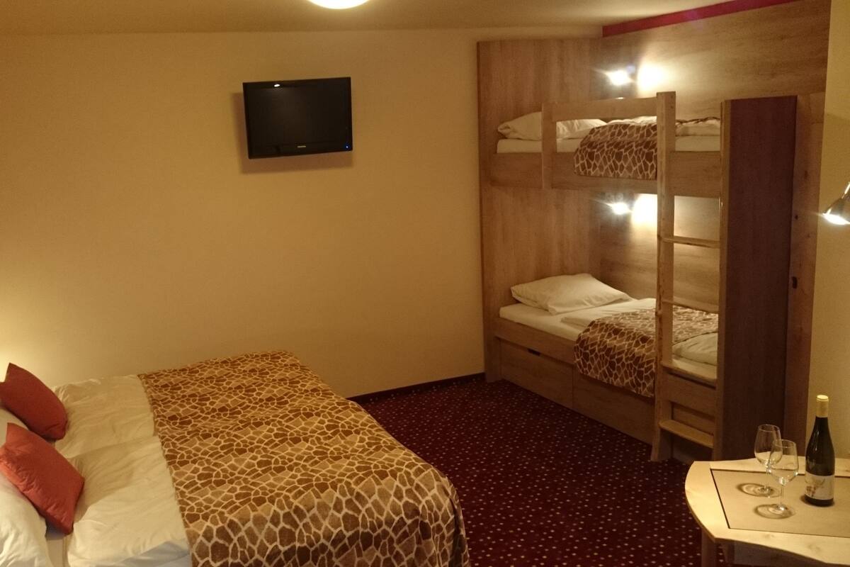 A hotel room with a single bed, a bunk bed, a TV mounted on the wall, a small table with glasses and a bottle, and a lamp on the left side.