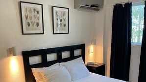 Un lit bien fait, avec une literie blanche, une tête de lit sombre et une couverture matelassée. Deux reproductions botaniques encadrées ornent le mur, ainsi qu’un ventilateur de plafond et un appareil de climatisation.