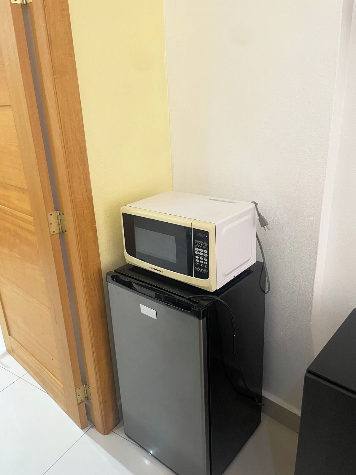 A compact refrigerator with a microwave on top, positioned against a wall with a door frame visible to the left.