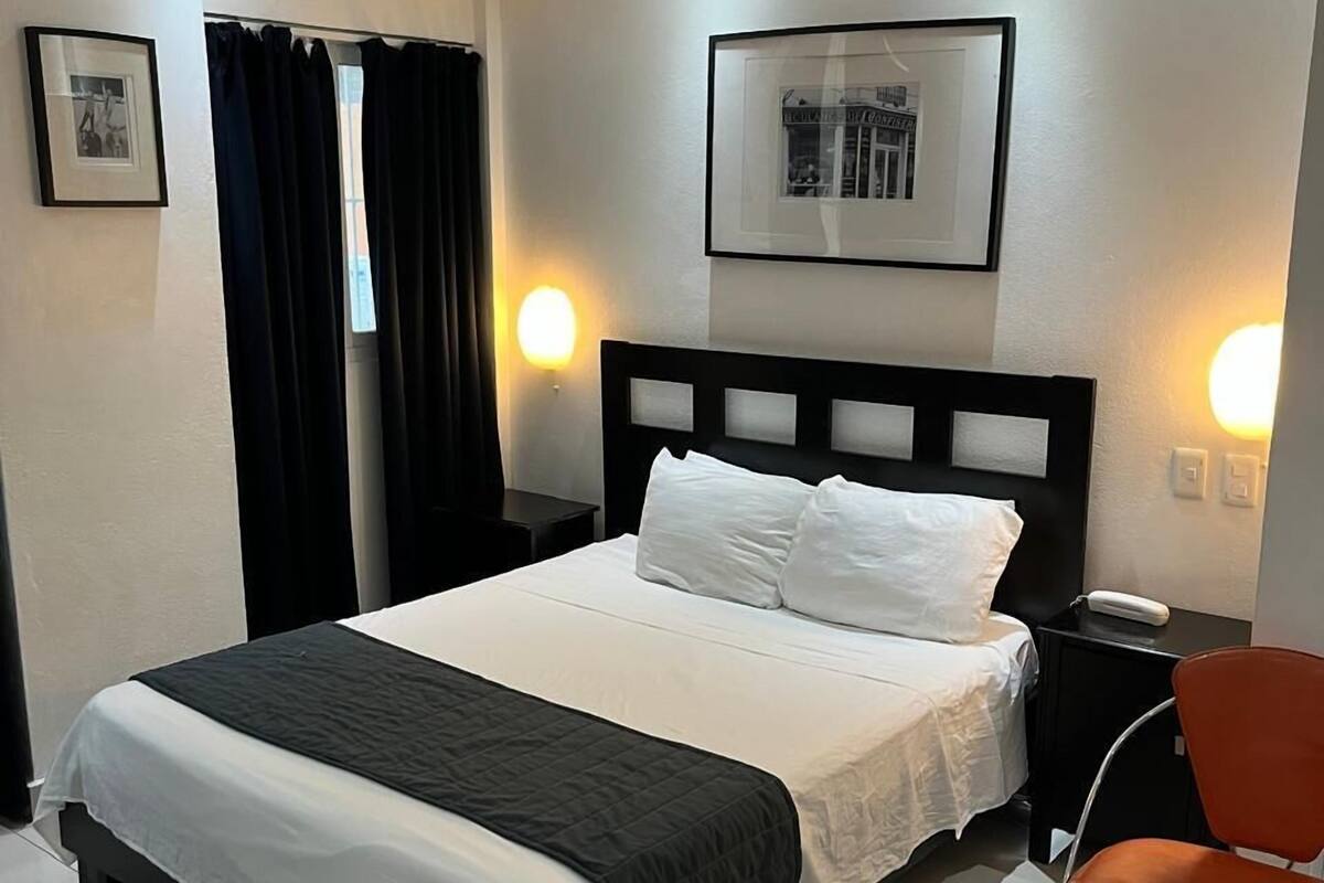 Ein ordentlich eingerichtetes Hotelzimmer mit einem Bett, einem Sessel, einem Nachttisch, einem Deckenventilator und einem gerahmten Bild an der Wand.