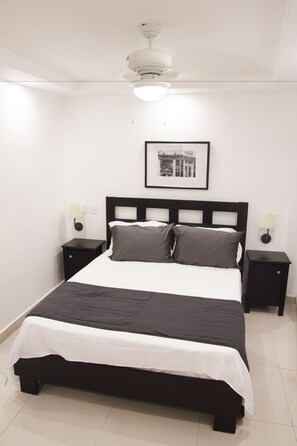 A neatly made bed with a black and white bedspread, two bedside tables with lamps, a ceiling fan, and a framed picture on the wall. - City Caribbean Hotel Boutique (Santo Domingo)