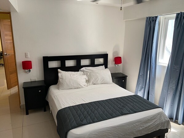 A neatly arranged bedroom with a bed, two bedside tables, a red wall lamp, and a window with curtains.