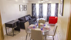 Un coin repas avec une table en verre dressée pour quatre personnes, entourée de chaises. On aperçoit un canapé noir et un fauteuil rouge à l’arrière-plan.