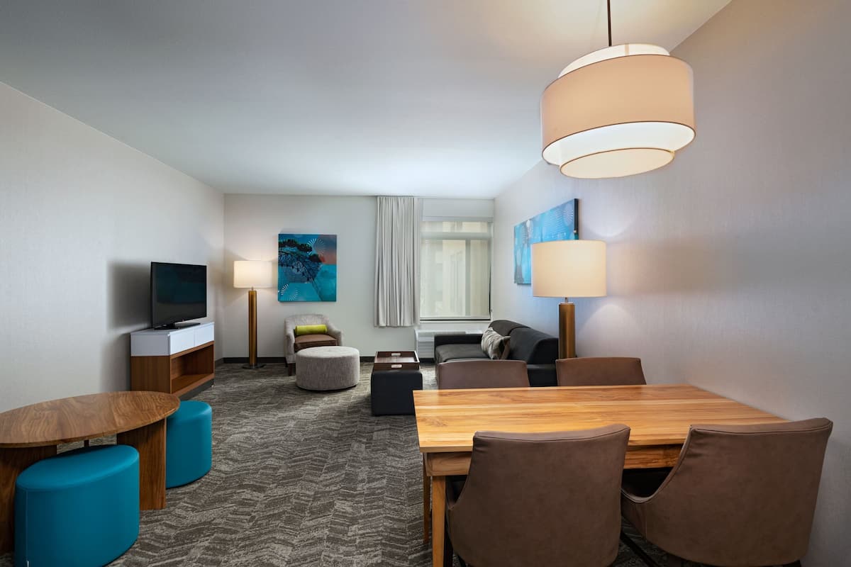 A hotel room with a wooden dining table, blue ottomans, a flat-screen TV, a sofa, a coffee table, and a large window with curtains.