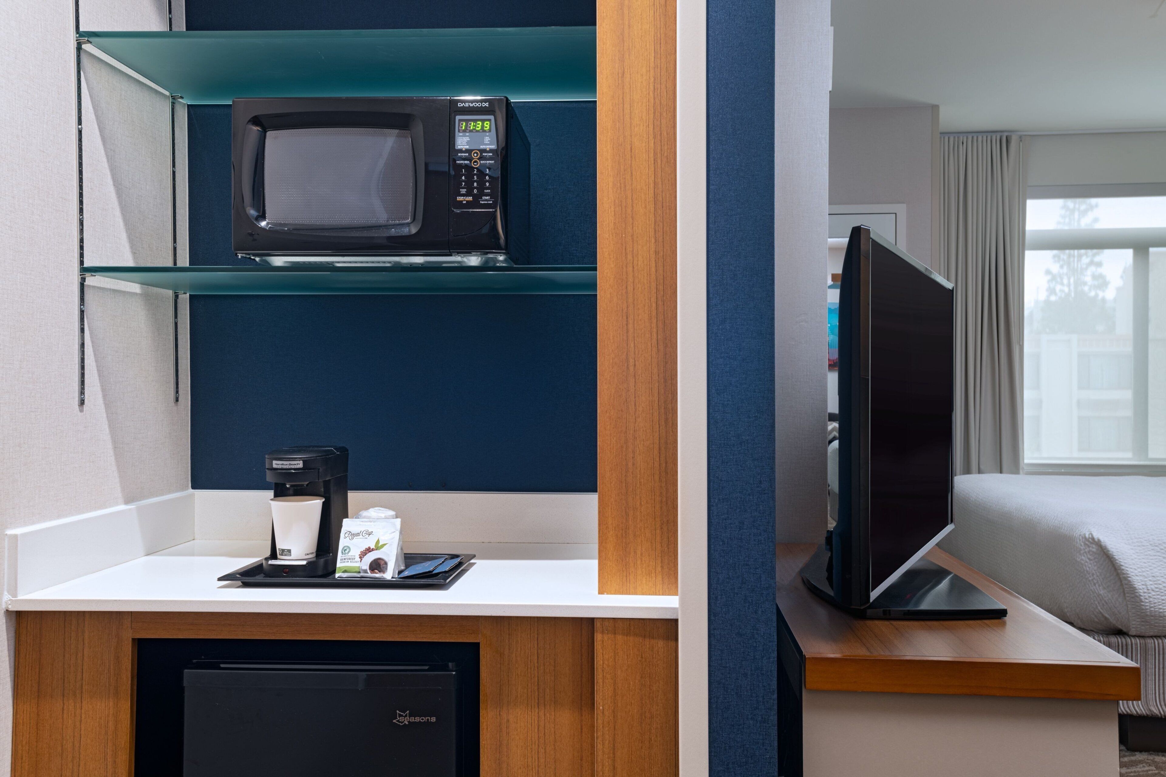 A hotel room with a microwave on a shelf, a coffee maker, a flat-screen TV, and a bed.
