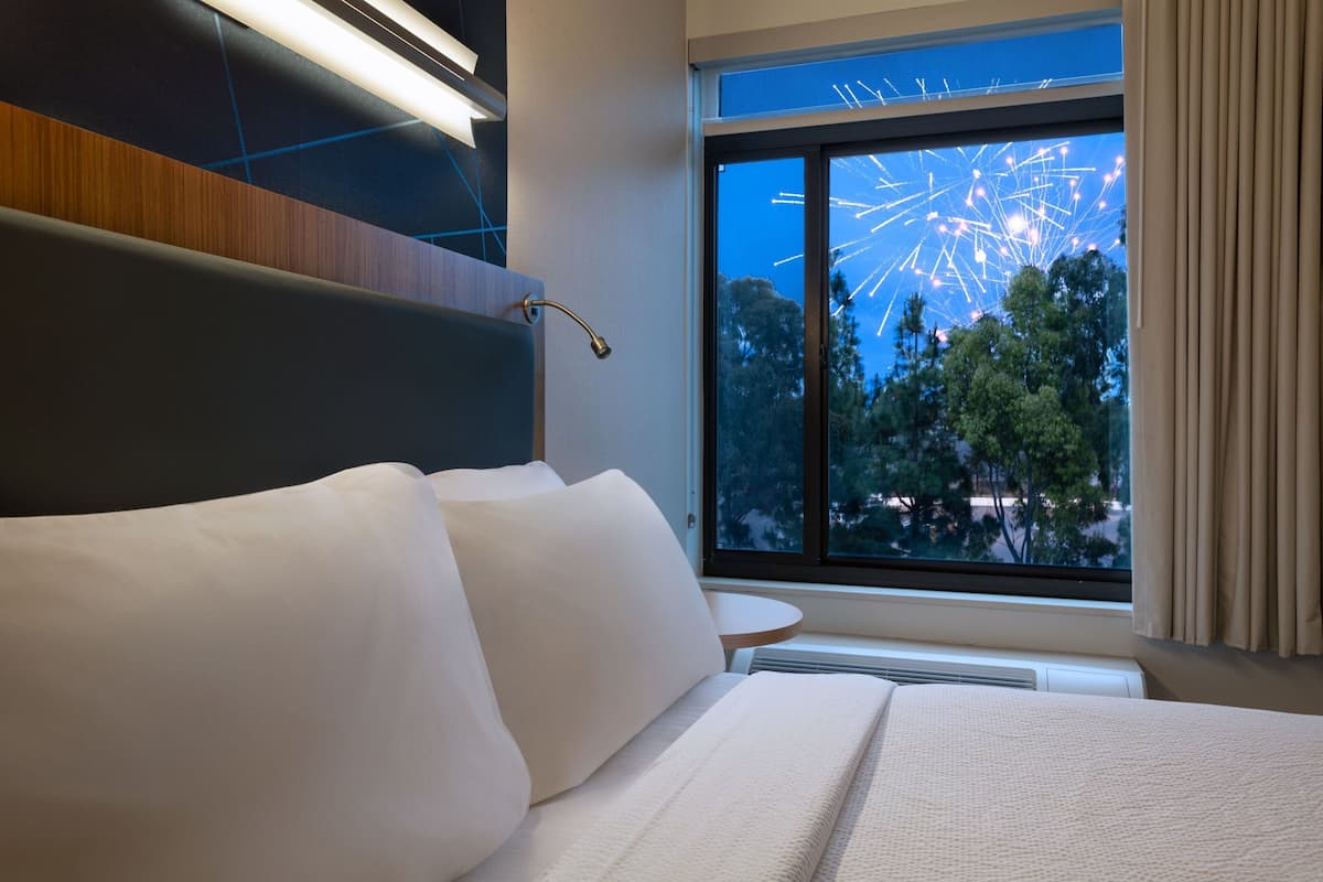 A hotel room with a large bed, a nightstand, a modern headboard, a window with a view of a Ferris wheel, and a ceiling light fixture.