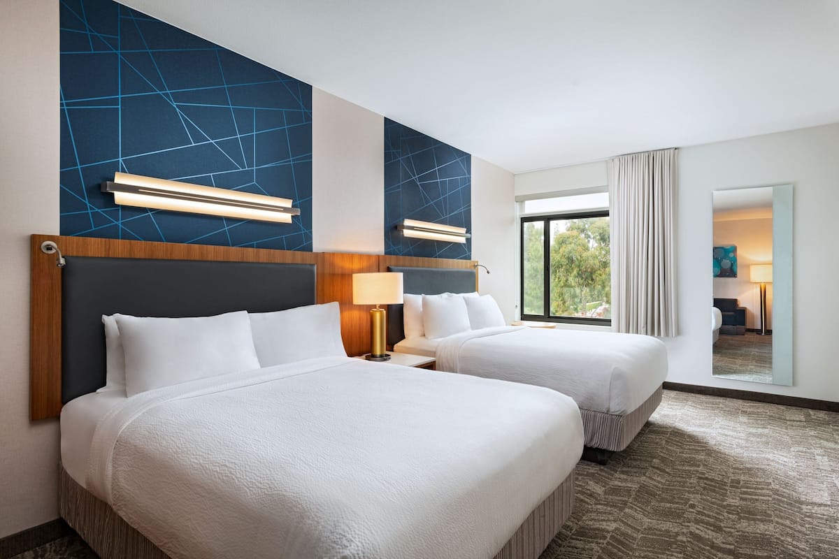 A hotel room with two beds, a large window, and a view of greenery outside.