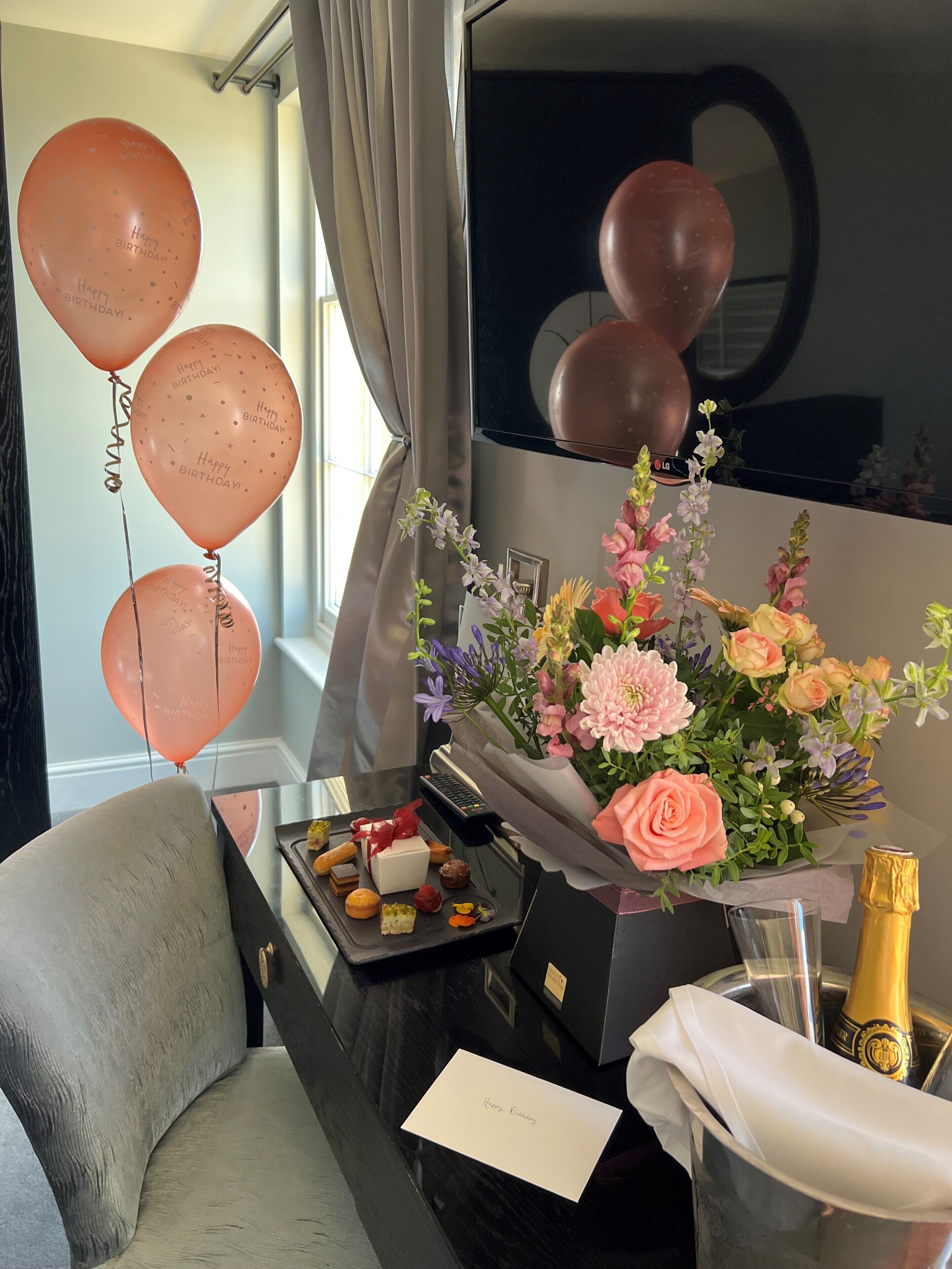 A hotel room with a desk, a bouquet of flowers, a bottle of champagne, and balloons.