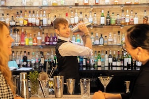 Um bar com um bartender preparando uma bebida, prateleiras com diversas garrafas de bebidas e utensílios de bar no balcão.