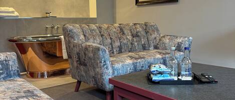 A modern living room with a patterned sofa, a wooden side table, a framed picture on the wall, and a small table with a remote control and bottles.