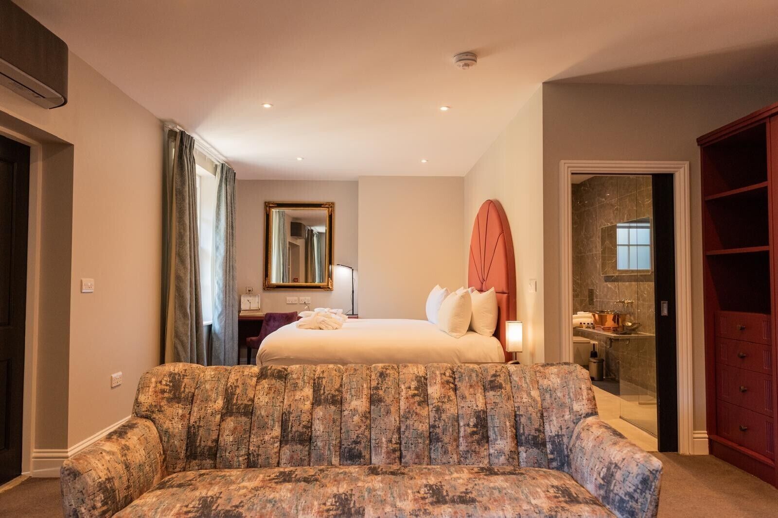 A hotel room with a bed, a sofa, a mirror, a bathroom with a shower, and a wardrobe.
