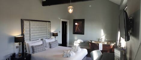 A hotel room with a large bed, bedside tables, a desk, a chair, a desk lamp, a hanging light fixture, a framed picture, and a mirror.