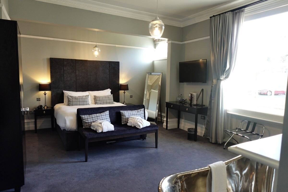 Habitación de hotel con cama, mesitas de noche, escritorio, silla, televisor y ventana con cortinas.