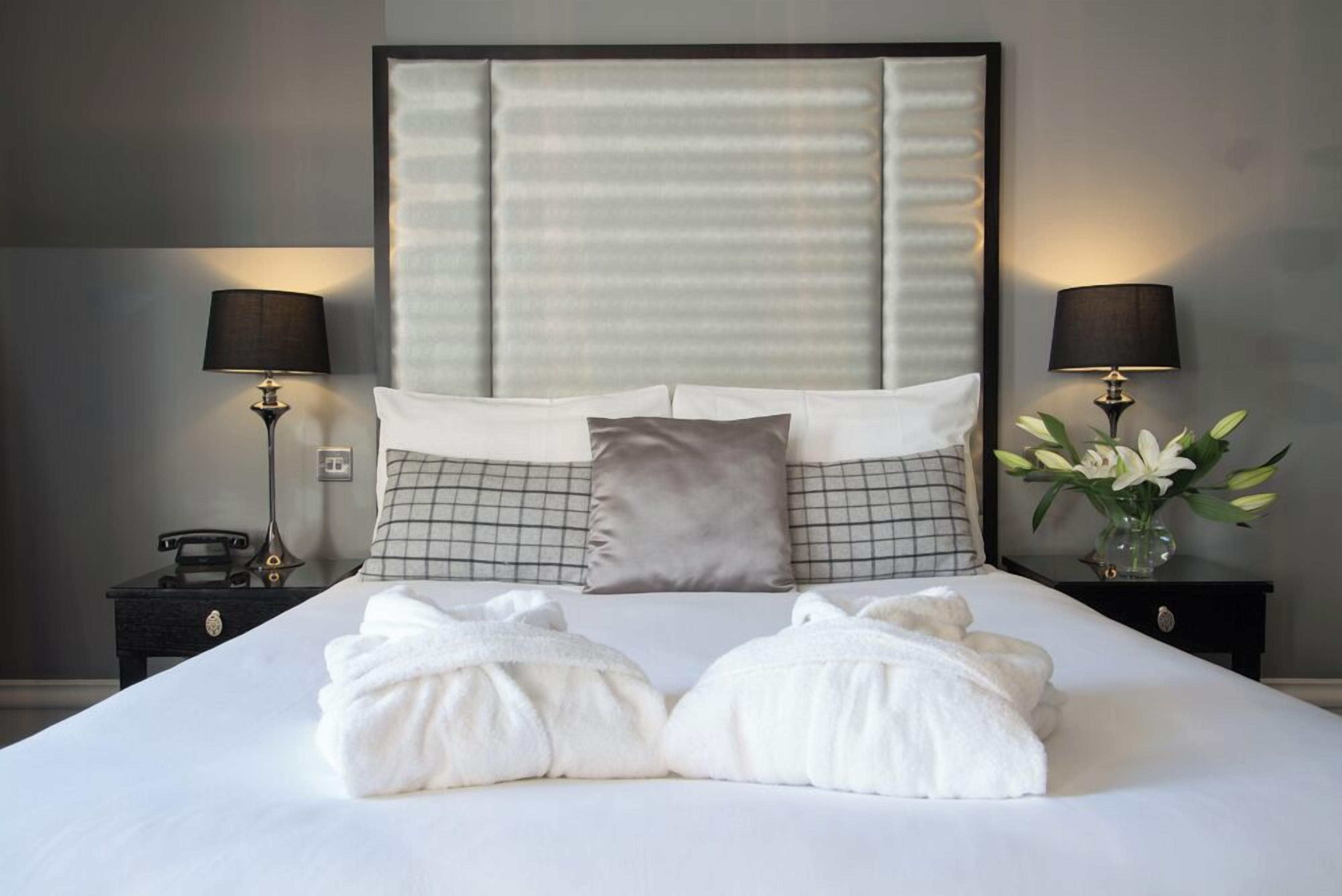 A neatly made bed with white linens, two bedside lamps, a phone on a nightstand, and a vase with flowers.