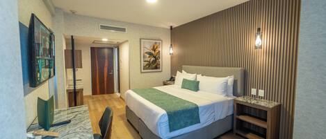 A hotel room with a bed, a desk, a chair, a nightstand, and a wall-mounted lamp.