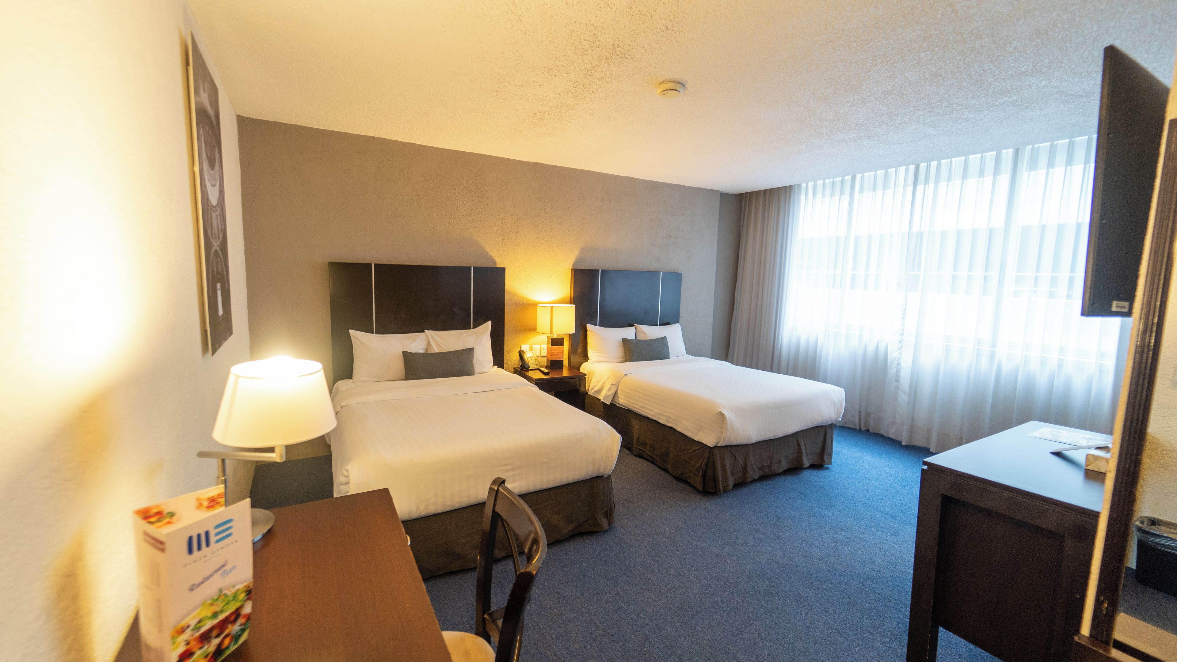 A hotel room with two beds, a desk, a chair, a lamp, and a window with curtains.