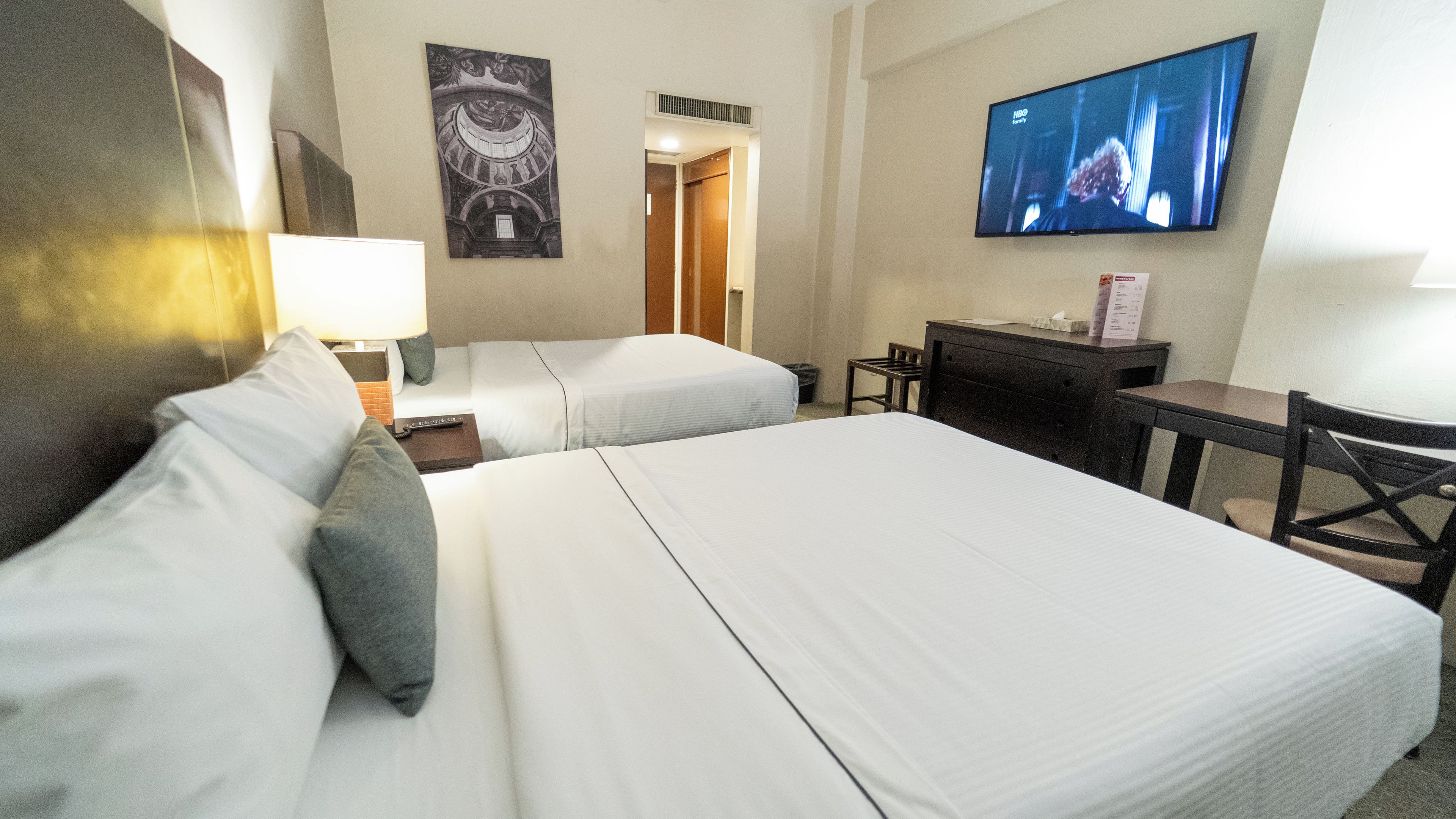 Une chambre d’hôtel avec deux lits, un bureau, une chaise, un téléviseur et une œuvre d’art accrochée au mur.