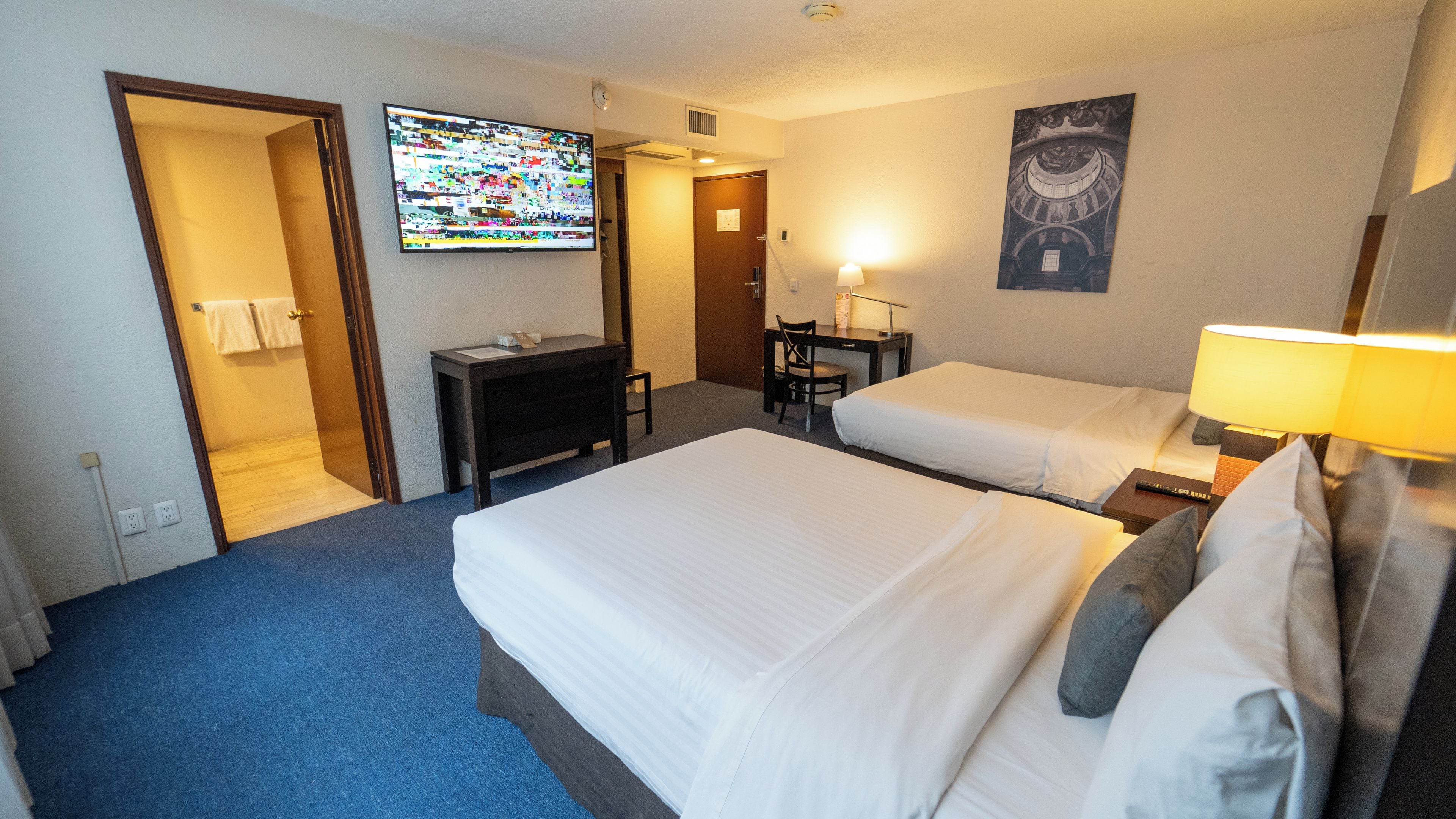 A hotel room with two beds, a desk, a chair, a television, and a painting on the wall.