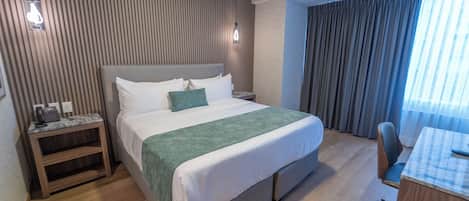 A hotel room with a bed, bedside table, desk, chair, and a view of the outside through a window.