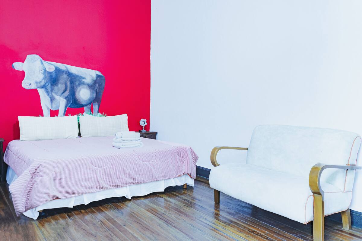 A bedroom with a bed, a white chair, and a red wall with a cow painting.