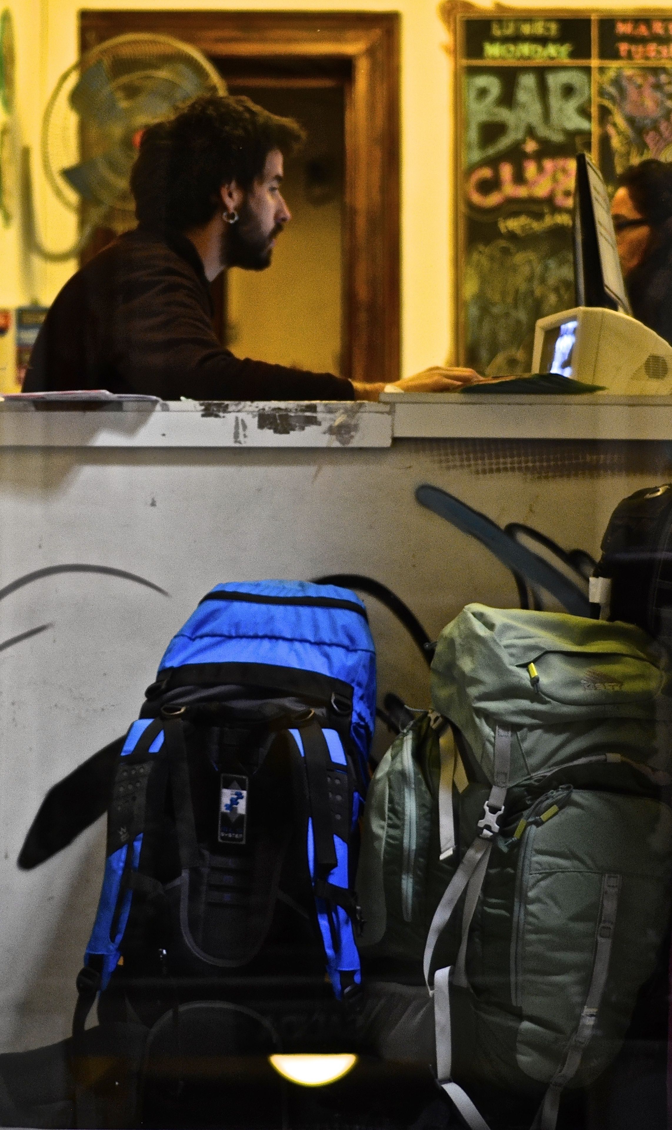 A person at a counter with a computer, surrounded by bags and a wall with graffiti.