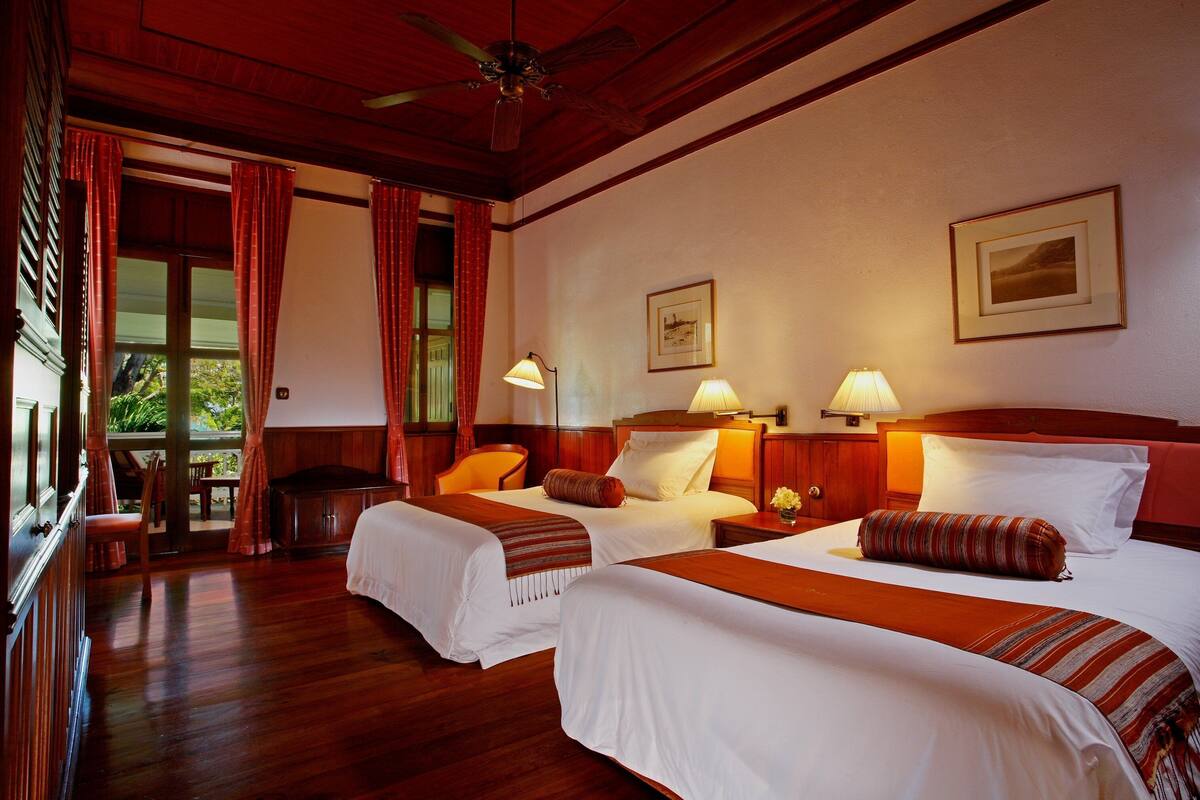 Ein Hotelzimmer mit zwei Betten, Holzboden, Deckenventilator und Blick durch ein Fenster mit roten Vorhängen.