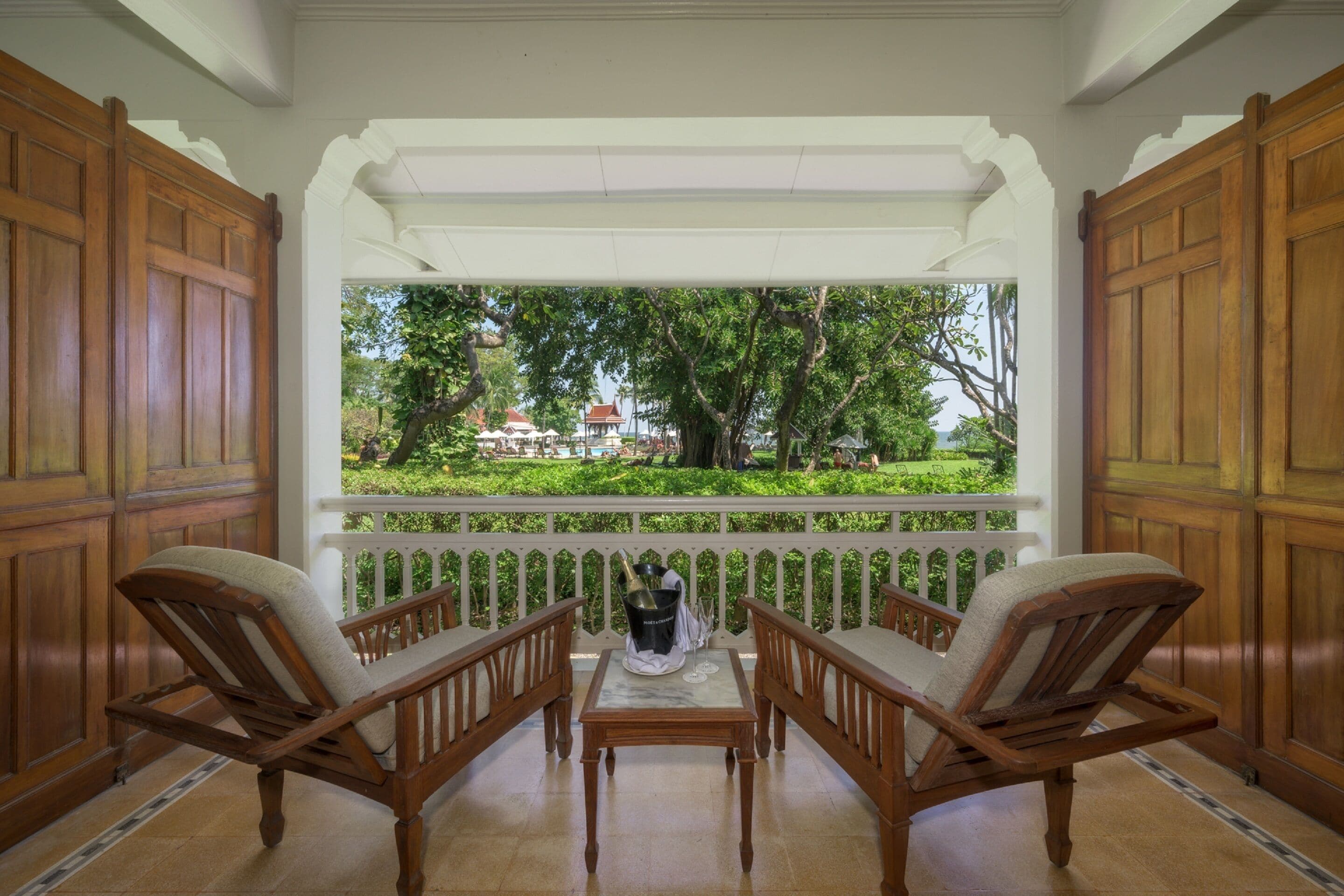 A wooden-framed porch with two chairs and a table, overlooking a lush garden and a building in the distance.