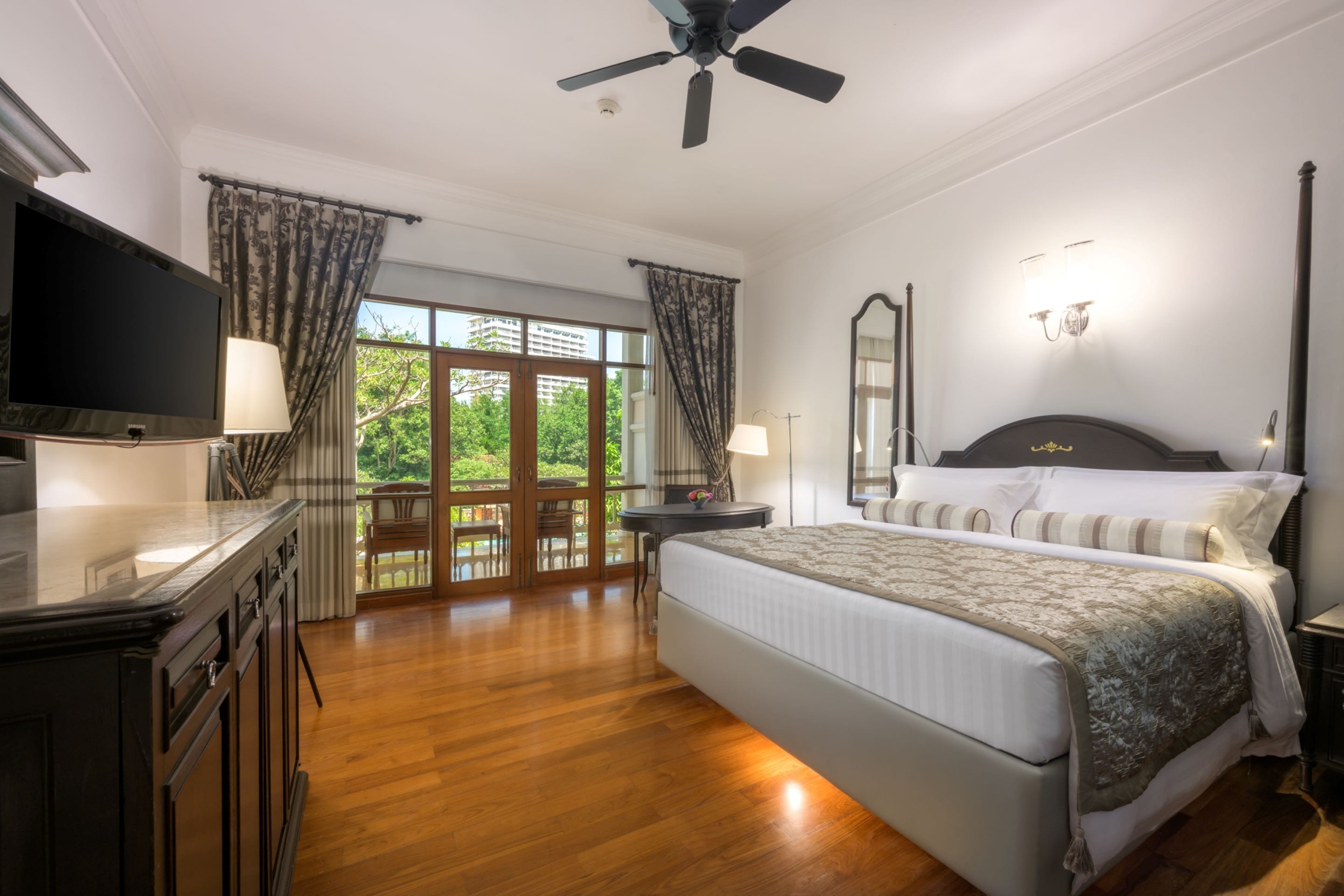 Ein geräumiges Schlafzimmer mit einem großen Bett, einem Flachbildfernseher, einem Deckenventilator und einem Balkon mit Blick auf Grünflächen.