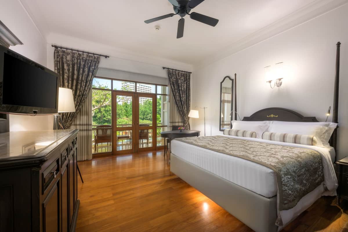 A spacious bedroom with a large bed, a flat-screen TV, a ceiling fan, and a balcony with a view of greenery.
