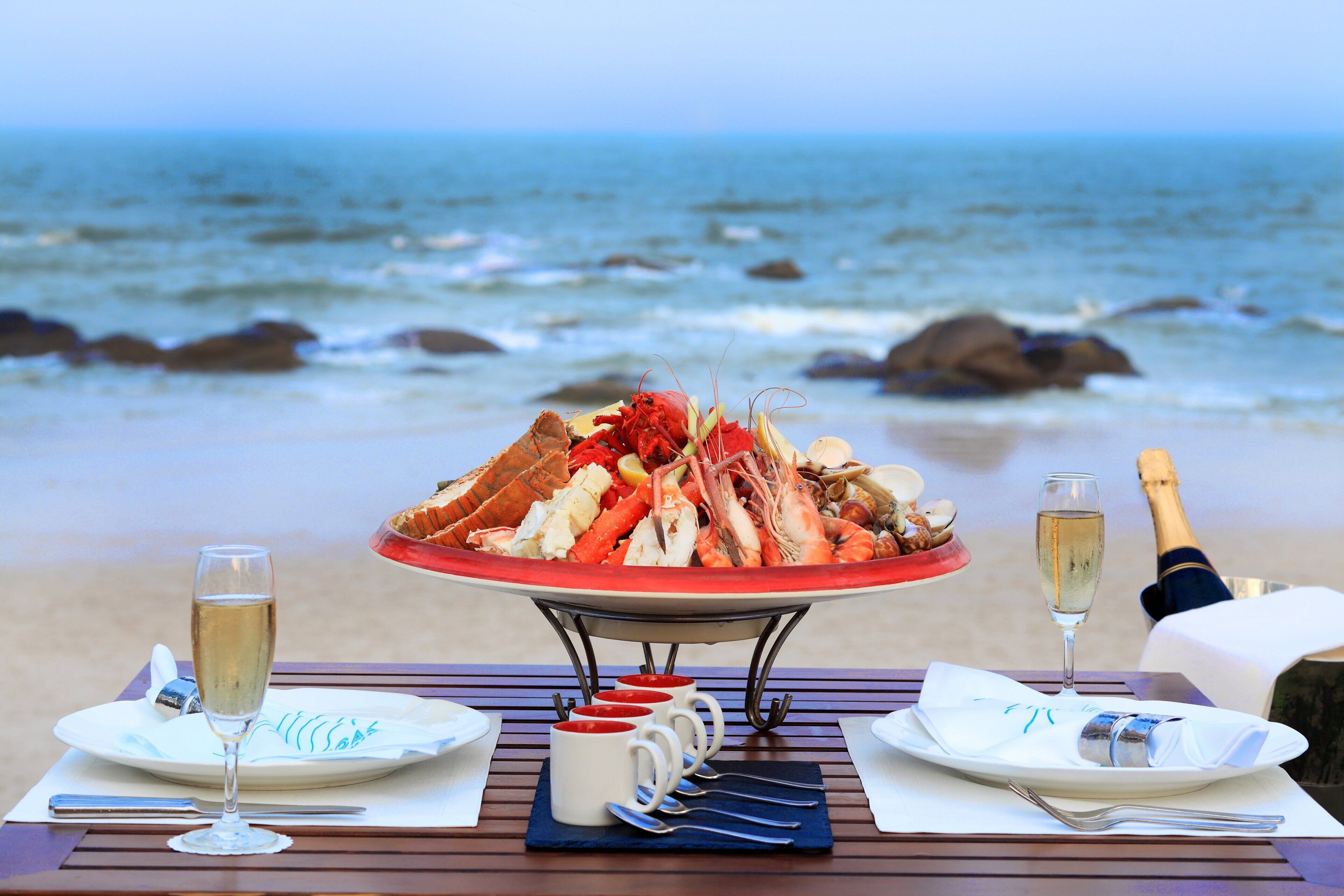 Un plato de mariscos sobre una mesa con una botella de champán y copas de vino, ubicado en una playa con el océano al fondo.