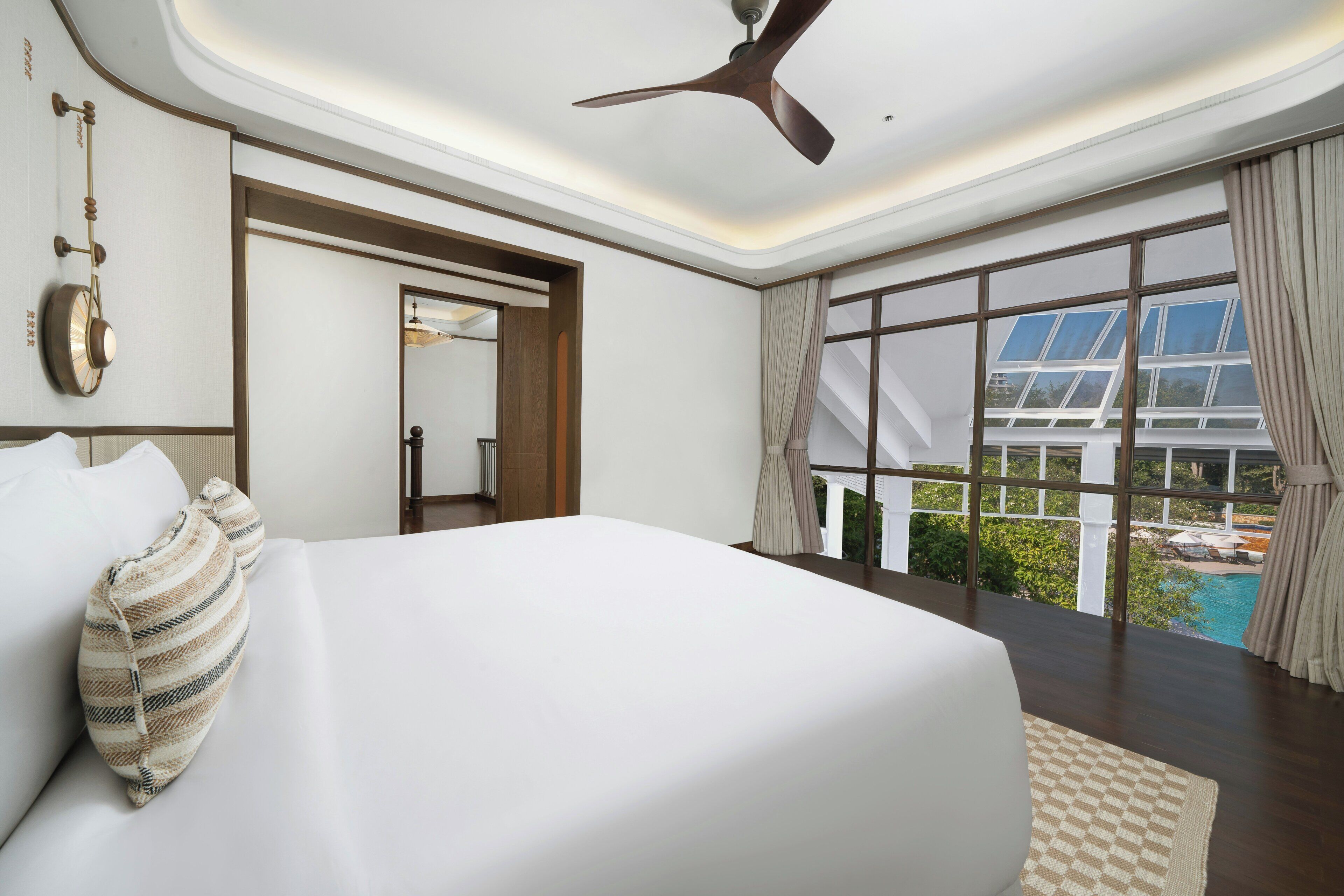 A modern hotel room with a large bed, a ceiling fan, a large window with a view of a swimming pool, and a wooden door.
