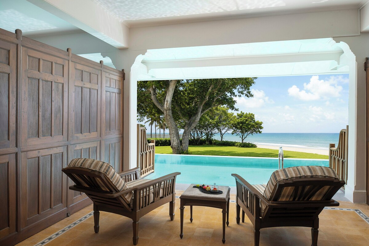 A room with a view of a pool and beach, featuring two wicker chairs and a small table.