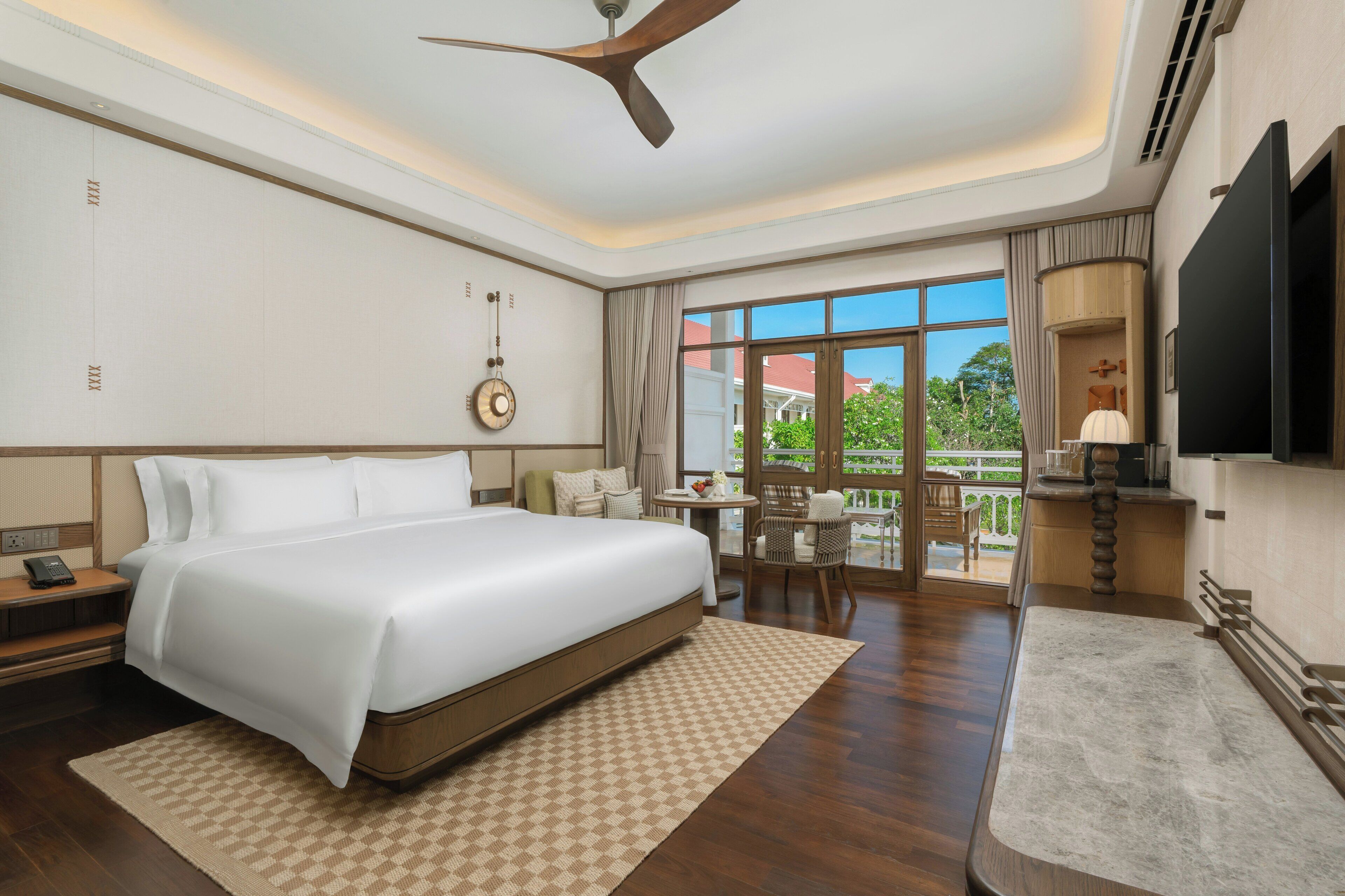 Ein Hotelzimmer mit einem großen Bett, einem Deckenventilator, einem Fernseher, einer Essecke mit Tisch und Stühlen sowie Blick ins Freie.