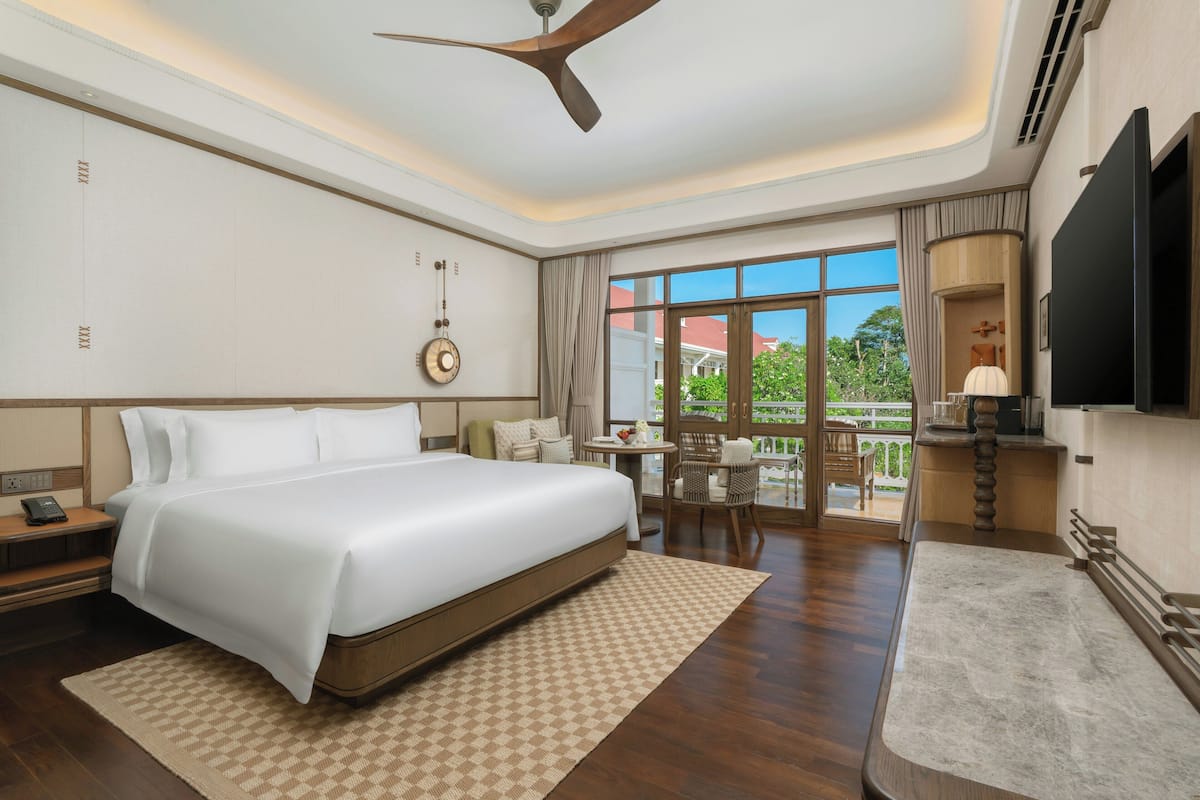A hotel room with a large bed, a ceiling fan, a TV, a dining area with a table and chairs, and a view of the outdoors.
