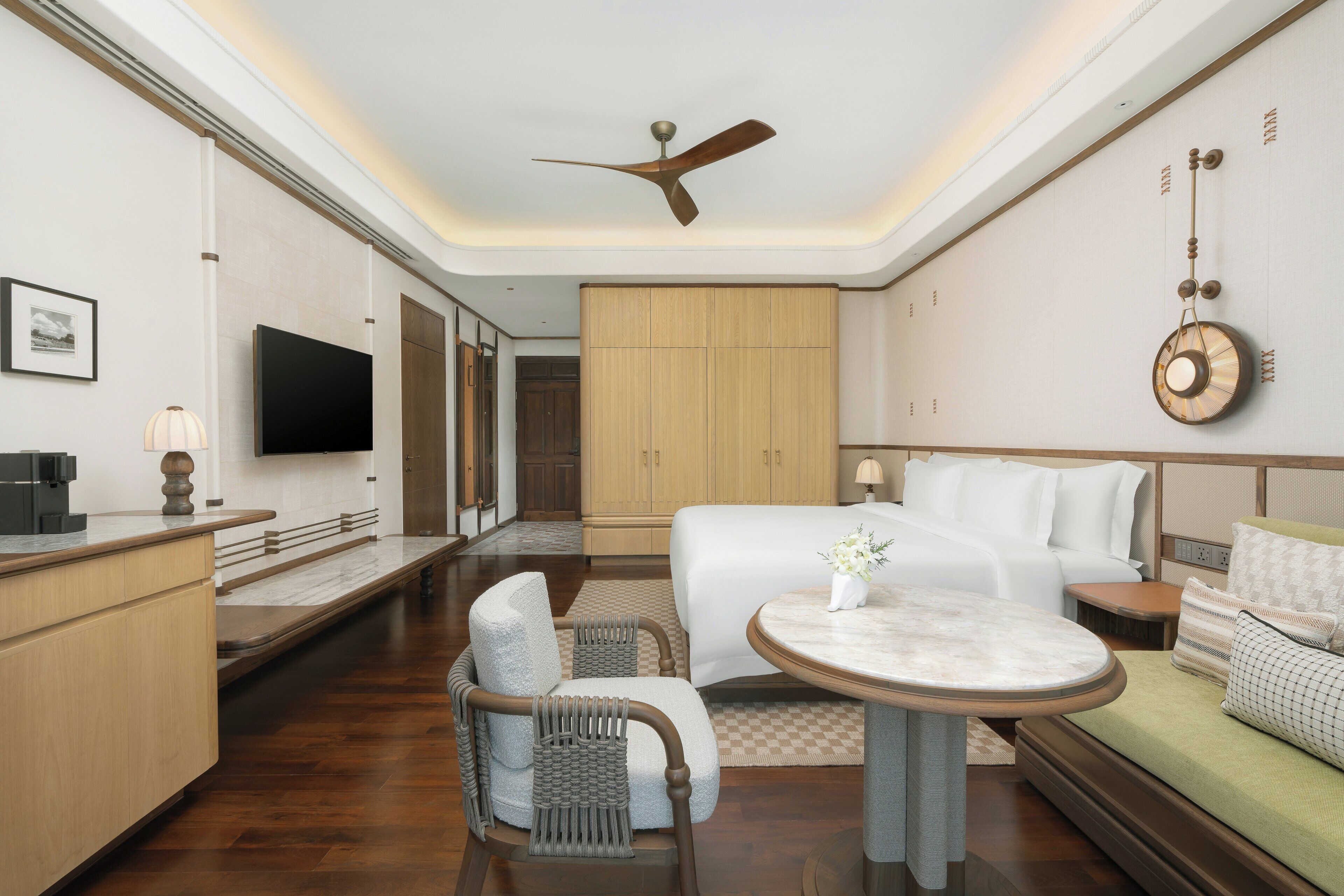 A hotel room with a bed, a TV, a desk, a chair, a table, and a ceiling fan.