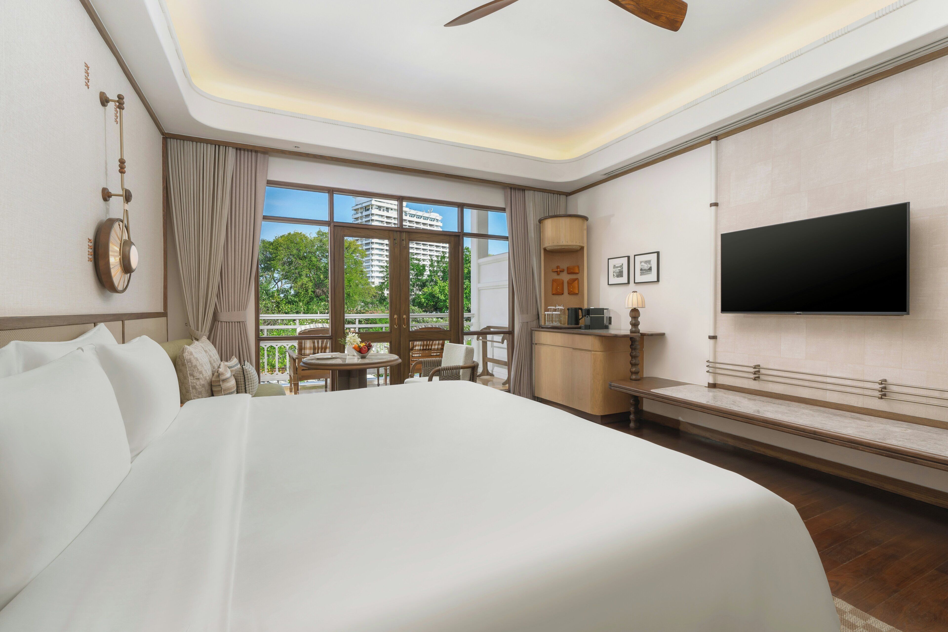 Une chambre d’hôtel avec un grand lit, une télévision, un coin repas avec une table et des chaises, et une vue sur les bâtiments par la fenêtre.