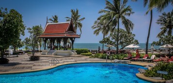 Zona de piscina con vista al mar, palmeras y un edificio tradicional.