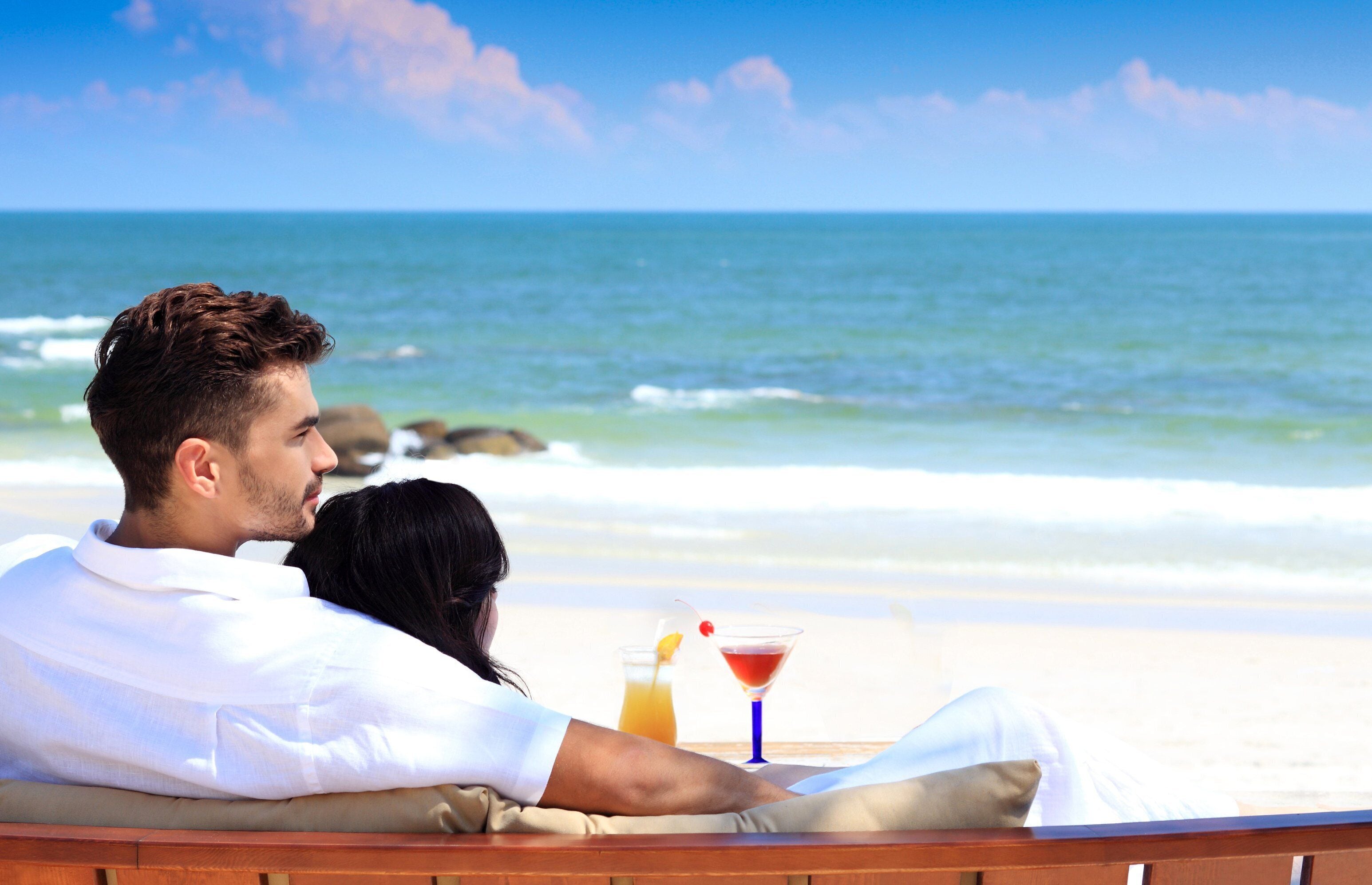 A couple relaxing on a wooden bench with a view of the ocean and a drink on the table.