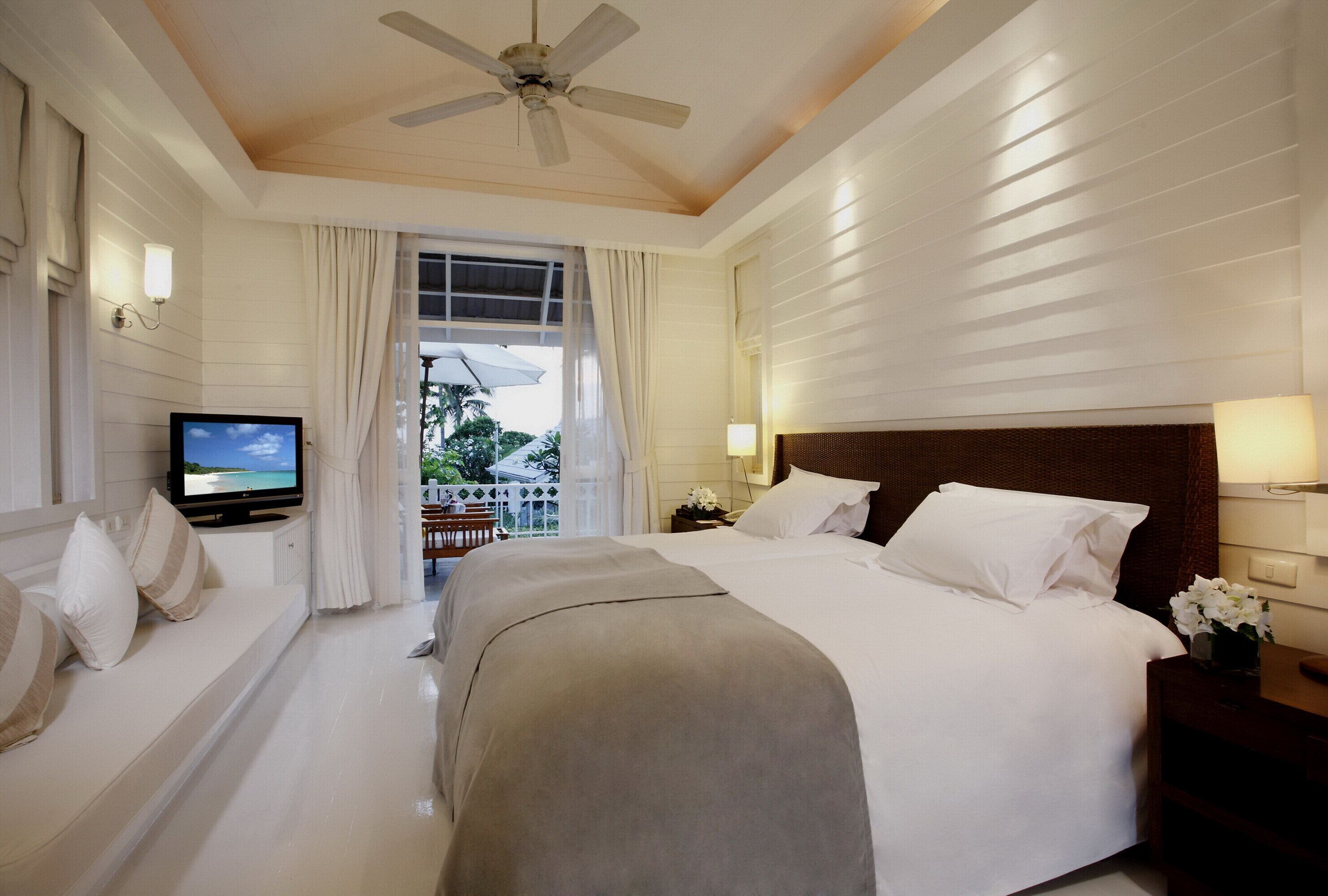 A hotel room with a bed, a television, a ceiling fan, and a view of the outdoors.