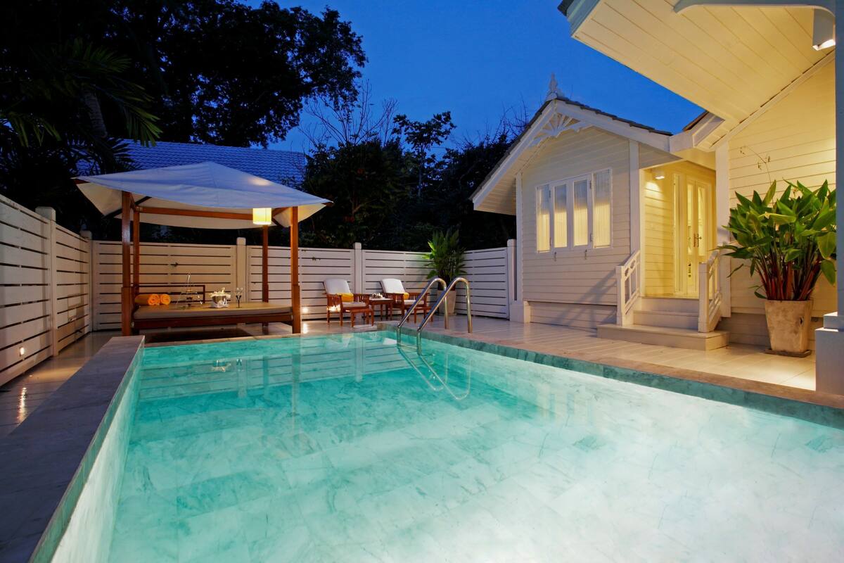 Ein Schwimmbecken mit überdachtem Sitzbereich, ein kleines Gebäude mit Veranda und ein Topfpflanze.