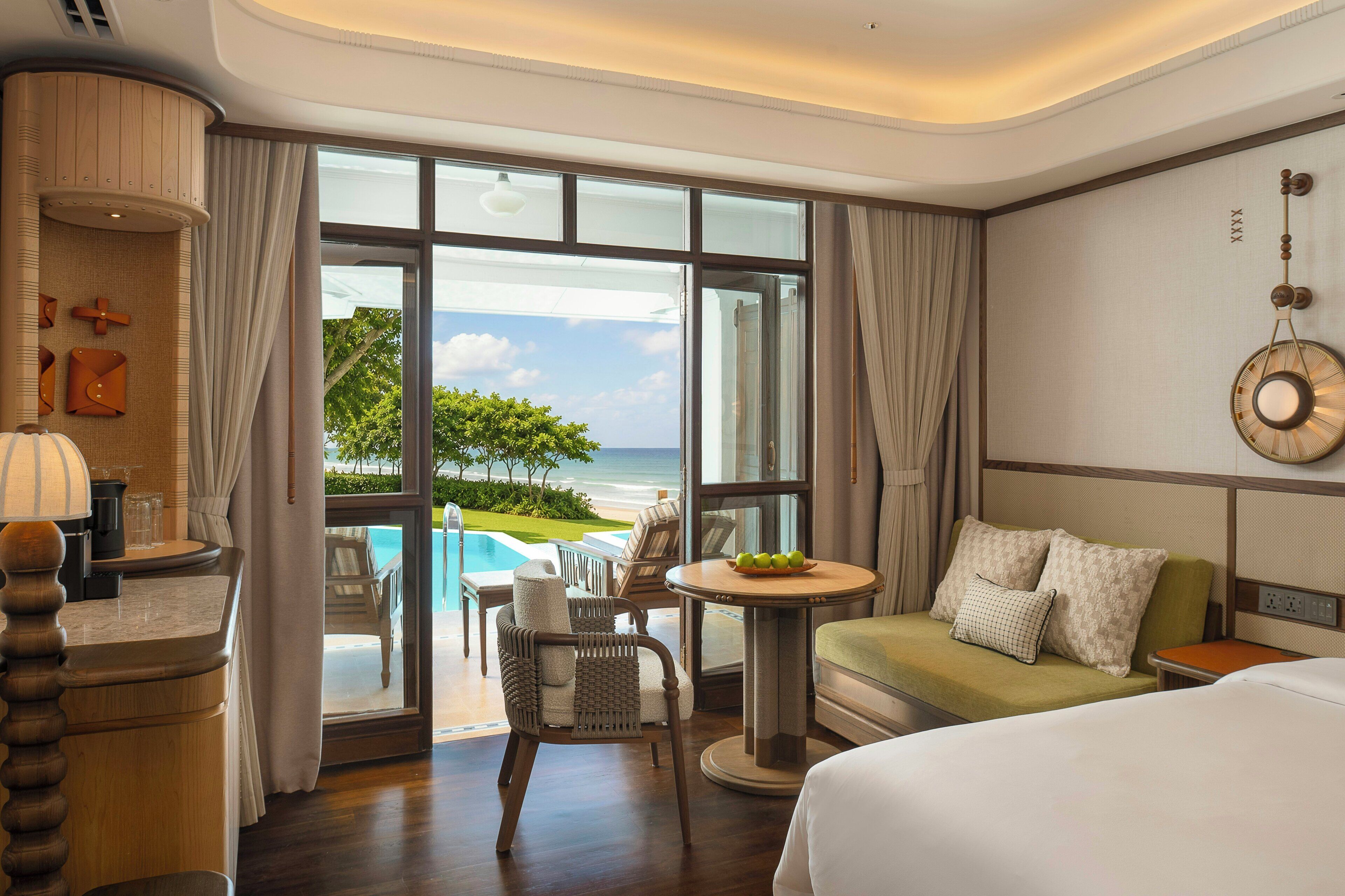 A hotel room with a large window, a bed, a round table with a bowl of fruit, a chair, and a view of the ocean.