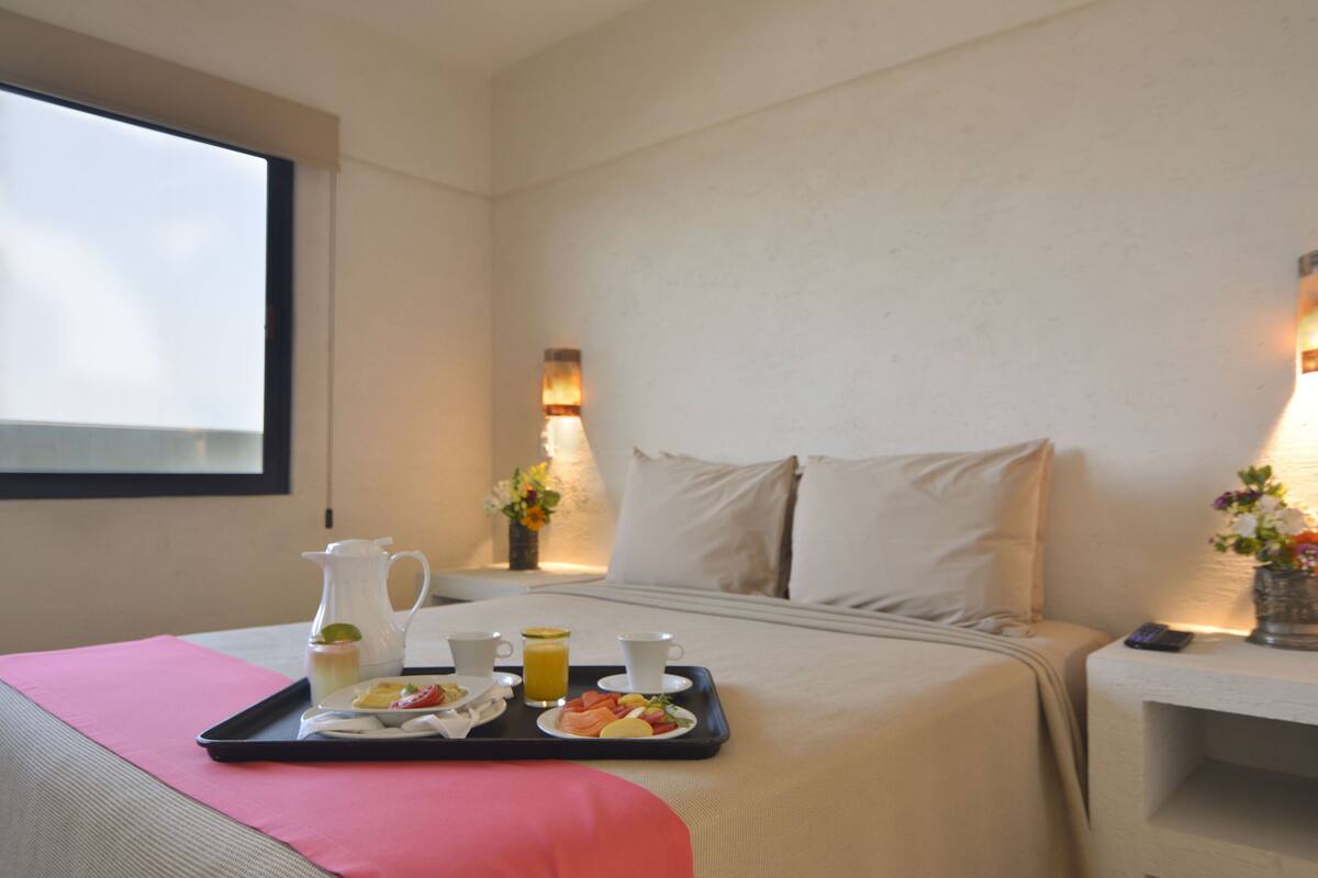 Ein Hotelzimmer mit einem ordentlich bezogenen Bett, einem Tablett mit Frühstücksgebäck, einer Karaffe und einer Vase mit Blumen.