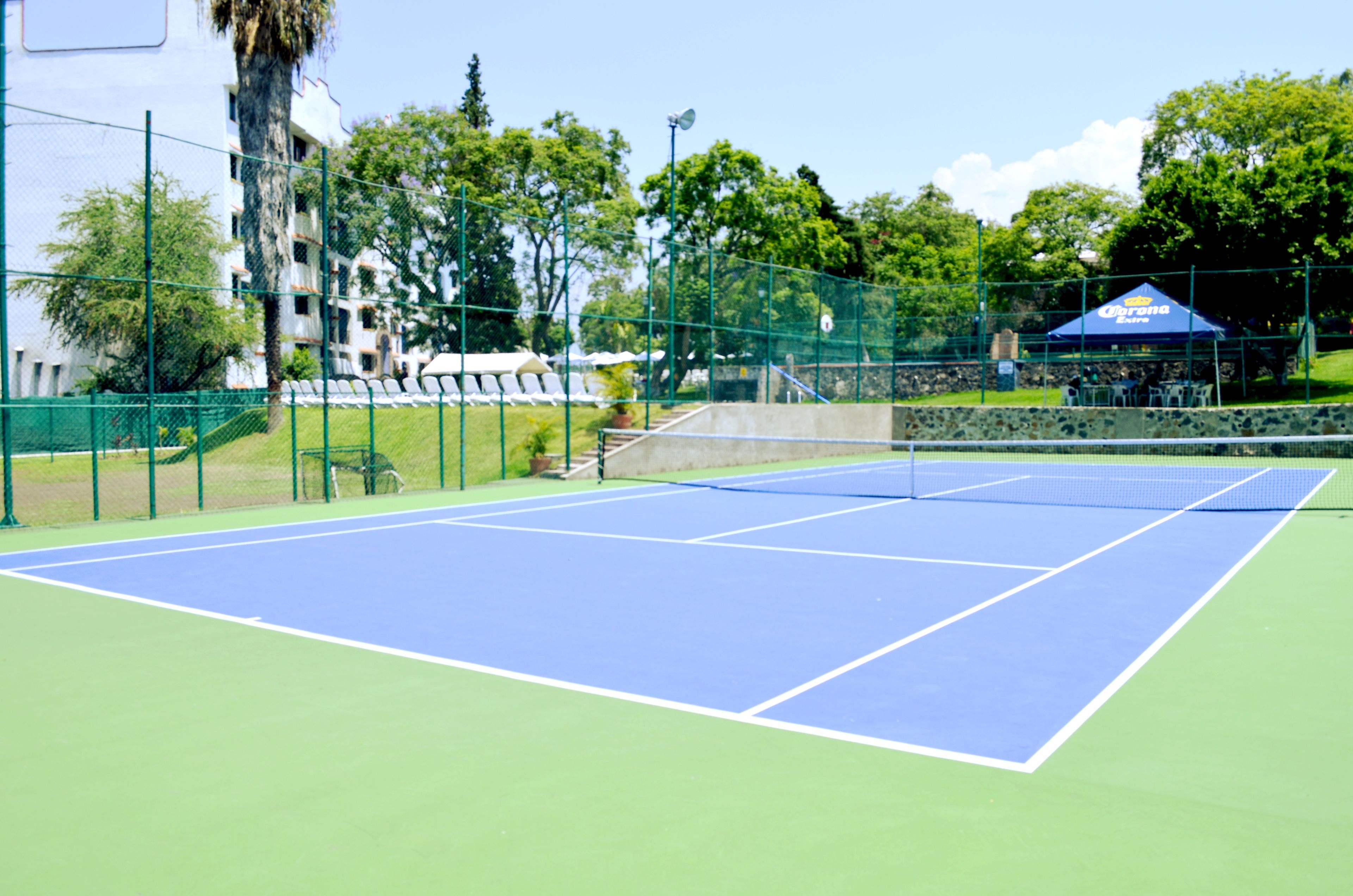 An outdoor tennis court with a blue playing surface, surrounded by a green fence, and adjacent to a building with a blue awning.